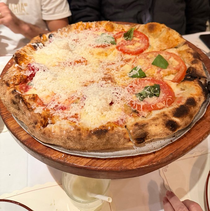 A freshly baked pizza with cheese, tomato slices, and basil served on a wooden stand at a restaurant table.