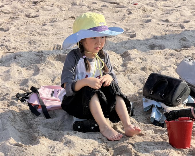 A young child in a colorful sun hat sits on the sandy beach with a pink life vest, black handbag, and red bucket nearby, appearing relaxed and looking off to the side.

The image doesn't relate to the post about a crypto KOL with major exchange partnerships—the photo shows a child at the beach, which seems unrelated or potentially ironic commentary compared to the high-stakes crypto promotion described.