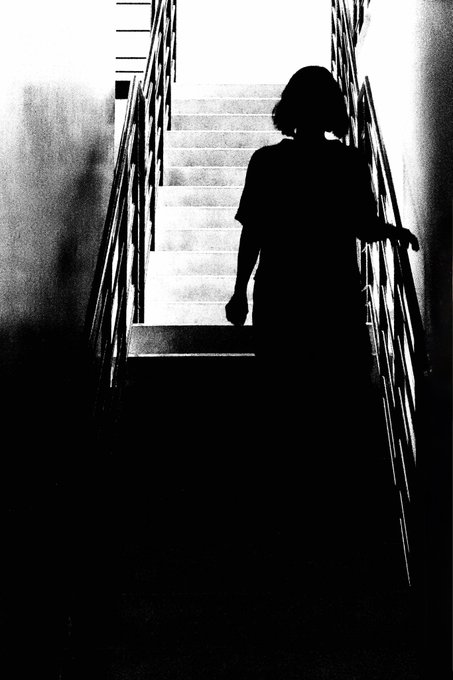 A high-contrast black and white photograph shows the silhouette of a person with short hair walking down a staircase toward a bright light.