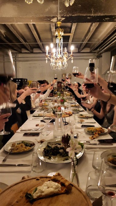 A dinner party scene with guests raising wine glasses in a toast around an elaborate chandelier-lit table filled with plates of food and floral centerpieces.