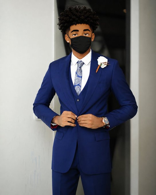 A young man with curly hair wears a blue three-piece suit with a patterned tie, white boutonniere, and a black face mask while buttoning his jacket.