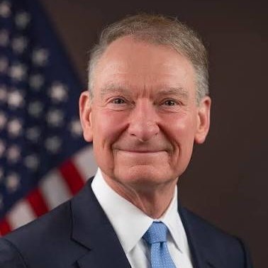 A professional portrait of an older man in a dark suit with a light blue tie, smiling, photographed in front of an American flag backdrop.