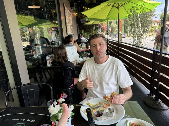 A man in a white t-shirt gives a thumbs up while holding a slice of pizza at an outdoor restaurant patio under green umbrellas, with a Zoberbacek laptop visible on the table.