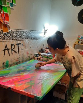 A young woman with her hair in a bun working on a colorful pink, green, and blue painted canvas in what appears to be an art studio with "ART" written on the wall.