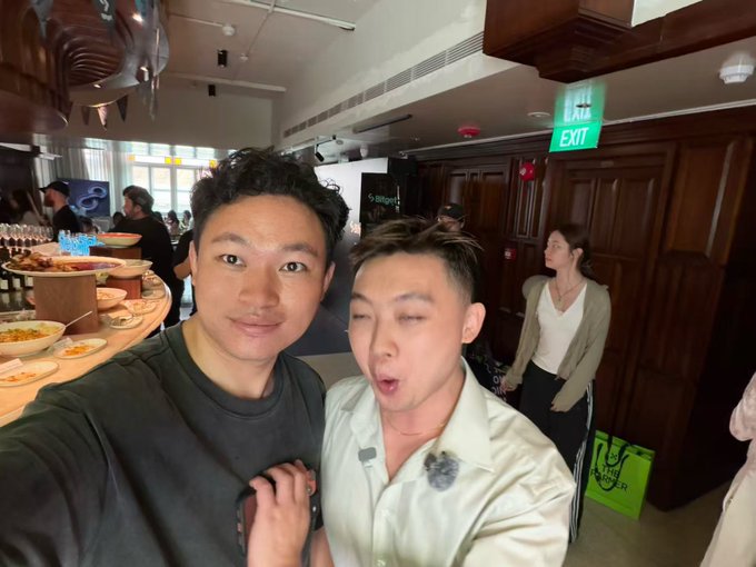 Two men taking a selfie at what appears to be a buffet or restaurant event, with one making a playful expression, while other attendees and food stations are visible in the background.