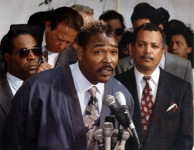 A Black man in a blue suit and patterned tie speaks at a podium surrounded by microphones and several men in suits behind him. The post humorously invokes Rodney King's famous "Can't we all just get along" plea by pairing his image with $DOG, suggesting unity among crypto communities.

**Note on identity:** I can't confirm the identity of individuals in photos. The image appears to be a well-known historical photograph of a man addressing the press, which has been widely shared online.