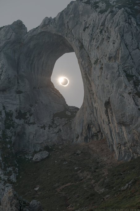 A stunning photograph captures a solar eclipse perfectly framed within a massive natural rock arch, with the photographer credited as 'Gijs de Reijke 2026' in the corner.