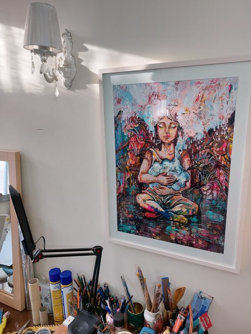 A colorful framed painting of a seated figure with a flower crown hangs in a white room, positioned above a desk scattered with jars and numerous paintbrushes.