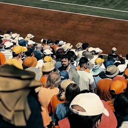 An overhead view shows a dense crowd of people seated in stands, many wearing hats, watching an event near a dirt arena with a green floor and red boundary.