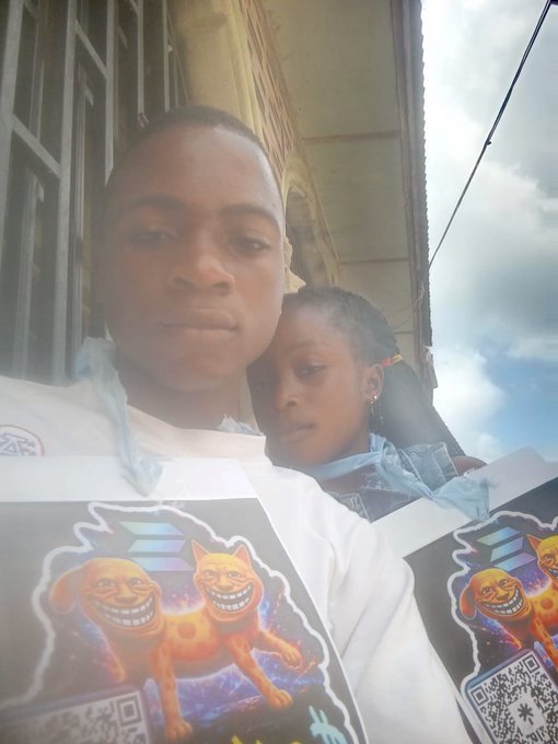 A selfie of two people wearing matching white t-shirts featuring grinning dog and cat memes with QR codes, taken outdoors.