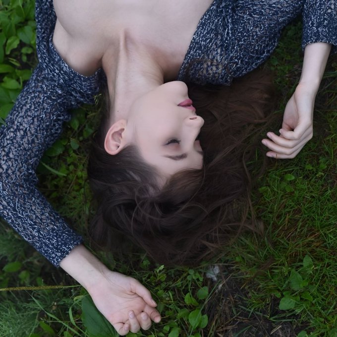 An overhead photograph of a woman with brown hair lying upside down on green grass, wearing a sheer blue knit top.