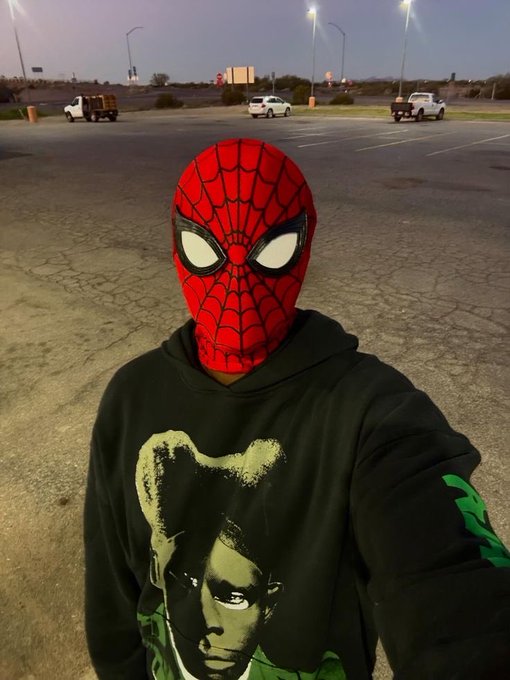 A selfie of a person wearing a red Spider-Man mask and a black hoodie featuring a green graphic, standing in a large empty parking lot at dusk.
