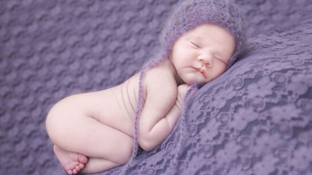 A sleeping newborn baby wearing a purple knitted hat rests peacefully on a textured purple blanket.