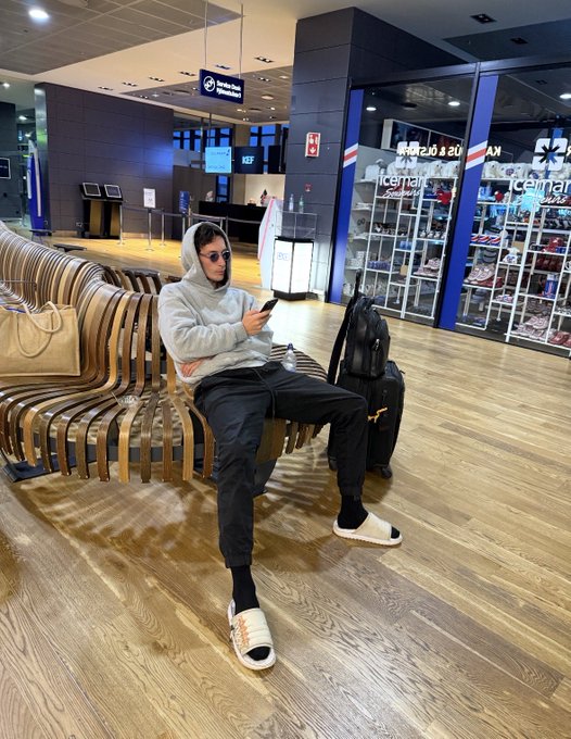 A young man in a gray hoodie and sunglasses relaxes on a wooden airport bench while scrolling on his phone, with an iceman express shop displaying "42-19-218 ROUTE 9 A.M." visible in the background. The image pairs with the bullish post about Litecoin (LTC), suggesting the trader is on the move yet staying locked into his crypto position, embodying the "close app" mindset.