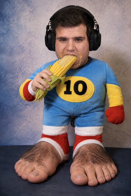 A creative composite photo of a man wearing headphones and a blue superhero costume with the number 10, eating corn on the cob, with grotesque enlarged feet edited into the image.