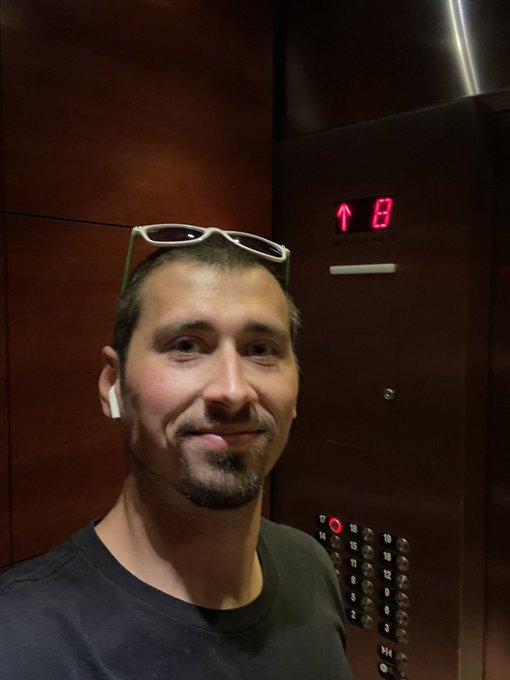 A man with short hair and sunglasses on his head takes a selfie inside an elevator showing floor buttons and an up arrow display.