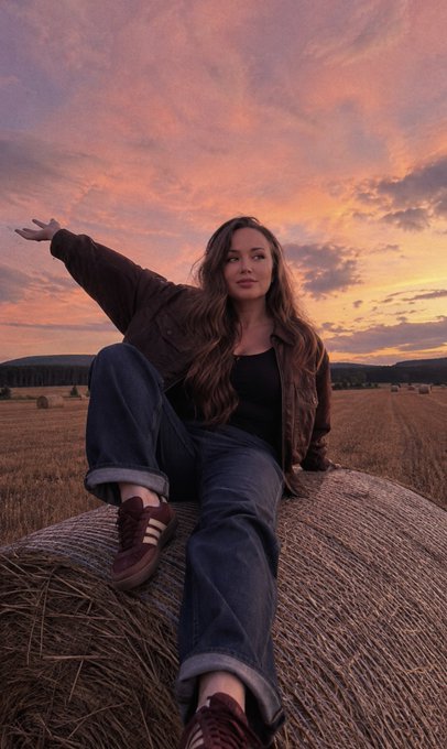 A young woman in jeans and a brown jacket sitting on a hay bale in a field at sunset, gesturing with one hand.