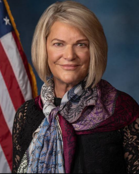 Senator Cynthia Lummis poses for an official portrait in front of an American flag, wearing a black lace top and a colorful purple and blue patterned scarf. The image accompanies a post about her urgent push for a Senate vote on the CLARITY Act, emphasizing her "now or never" stance on passing the crypto market structure legislation before Congress changes hands.