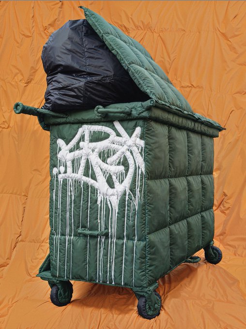 An olive green puffy down jacket displayed on a wheeled, cabinet-like structure with graffiti-style white paint dripping down the side, set against a wrinkled orange backdrop.