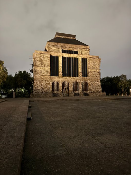 A photo of a large stone building with vertical window slits and a pyramid-shaped roof, viewed from a wide paved plaza under an overcast sky.