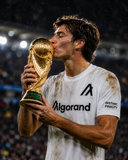 A man in a white Algorand-sponsored jersey kissing the FIFA World Cup trophy in a stadium, celebrating a championship victory.