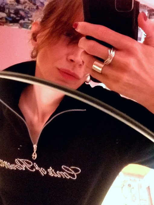 A selfie of a person taking a photo in a mirror, taking up most of the frame with a phone, wearing a black zip-up top with cursive text and several rings on their fingers.