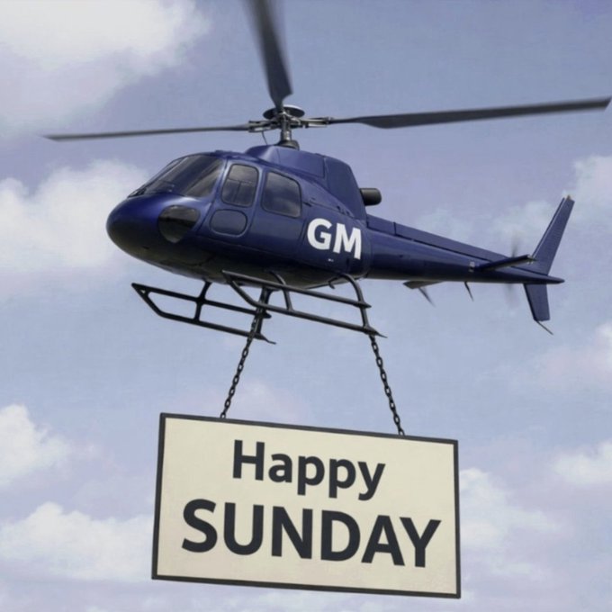 A dark blue helicopter labeled 'GM' flies in a partly cloudy sky, carrying a large banner suspended by chains that reads 'Happy SUNDAY'.