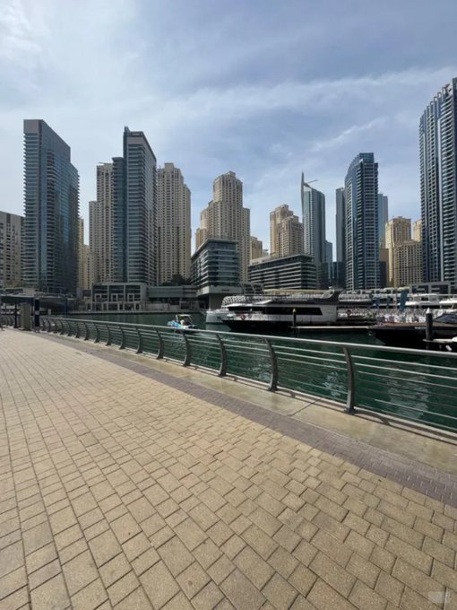 A scenic view of a modern city skyline with numerous high-rise buildings rising above a waterfront marina, seen from a paved walkway with metal railings.