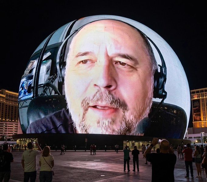 The Las Vegas Sphere displays a large image of a man wearing headphones, with people standing below watching the display at night.