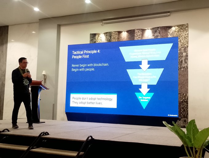 A man presents on stage in front of a large screen displaying 'Tactical Principle 4: People First — Never begin with blockchain. Begin with people.'