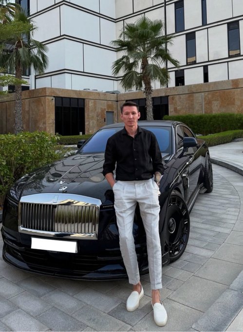 A man in a black shirt and white pants poses in front of a black Rolls-Royce luxury sedan parked outside a modern building with palm trees.