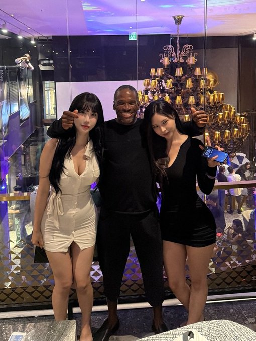 A man poses between two women in white and black dresses at what appears to be an upscale venue with a chandelier in the background.