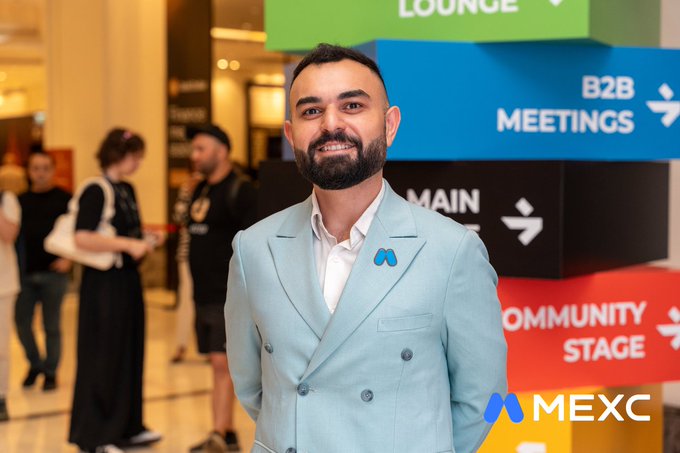 The image features a man wearing a light blue suit standing in front of a group of people who appear to be engaged in conversation around him. The man has a beard, and he is smiling at the camera. The background includes several people gathered together, some closer to the foreground while others are further back. There are also two large boxes visible in the scene, one on each side of the man's head.