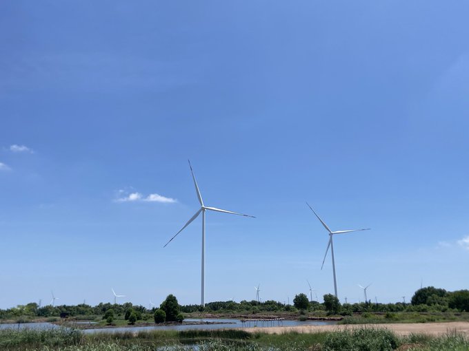 Landscape photograph of two large wind turbines in a green field with a body of water in the foreground under a bright blue sky with scattered clouds.
