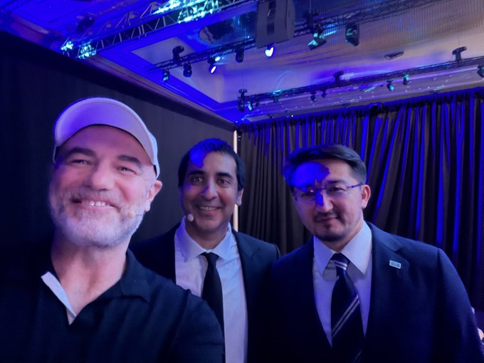 Three men pose together for a photo at an indoor event with blue stage lighting, including one in a white cap and two in suits with microphones.