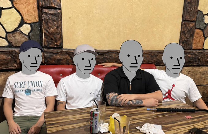 A photo of four people sitting at a wooden table in a restaurant with stone walls, their faces obscured by gray cartoon masks; one person wears a 'SURF UNION' t-shirt.