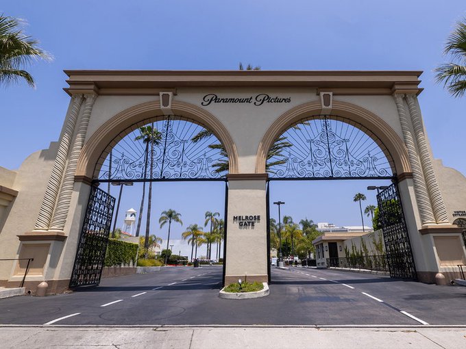The iconic twin arches of the Paramount Pictures studio entrance, marked 'Melrose Gate,' under a clear blue sky with palm trees in the background.