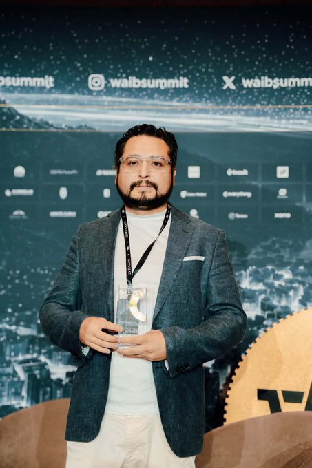 A man with glasses and a beard, wearing a dark blazer over a white shirt, holds a glass trophy at the WAIB Summit in Monaco, standing in front of a backdrop displaying the summit's social media handles (@waibsummit) and sponsor logos. The image aligns with the post celebrating Brickken's recognition as a "Rising Star" award recipient, visually capturing the moment of the achievement mentioned in the caption.