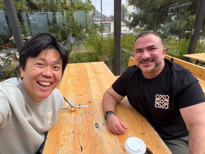 Two men smiling at the camera while sitting at an outdoor wooden picnic table, with a coffee cup on the table.
