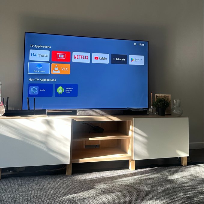 A television displaying a smart TV interface with TV Applications including TiViMate, Netflix, YouTube, VLC, and Google Play Store, sitting on a white and wood media console in a sunlit room.