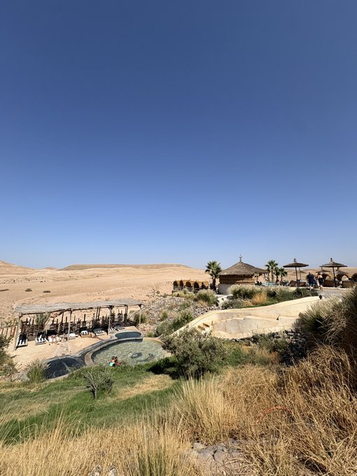 A scenic landscape featuring a desert resort with thatched-roof cabanas, a pool area with people, palm trees, and dry rolling hills under a clear blue sky.