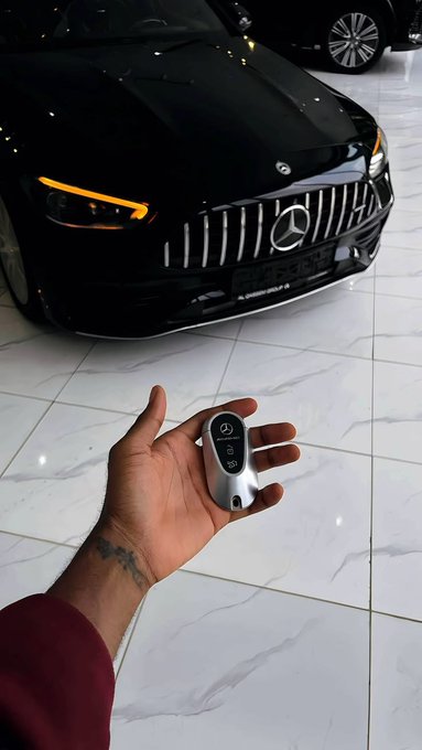 A first-person view shows a hand holding a Mercedes-Benz key fob in front of a sleek black Mercedes-AMG coupe parked on a white tiled floor.