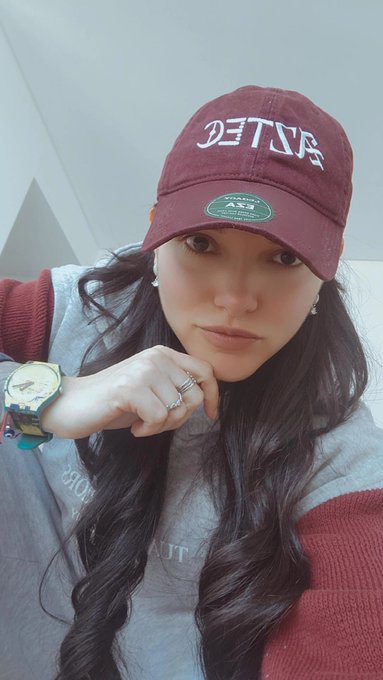 A selfie of a young woman wearing a maroon cap with mirrored text reading 'DETACH', a grey sweater over a red top, a green watch, and multiple rings.