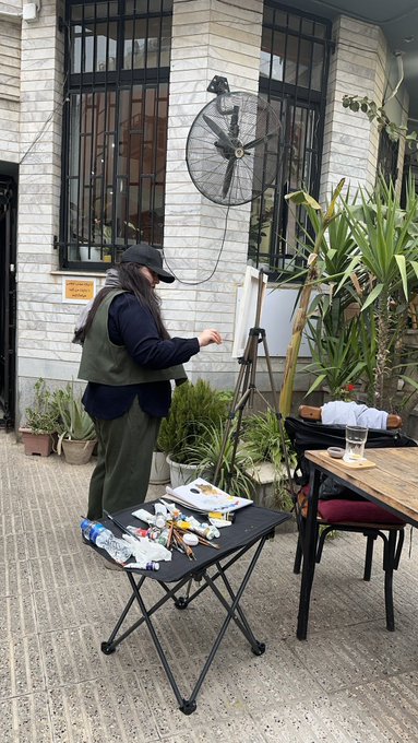 A bearded man in a green vest and black cap stands painting at an easel outside a white brick building, with art supplies arranged on a small folding table beside him.