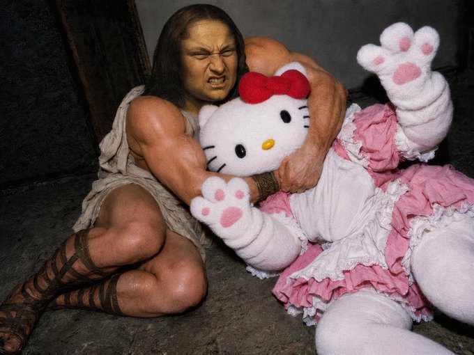 A cosplay or photoshoot image depicting a muscular figure dressed as Conan the Barbarian aggressively grappling with a large person in a Hello Kitty mascot costume on a stone floor.