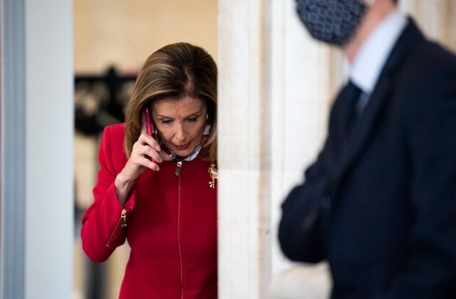 The image shows a woman wearing a red jacket talking on her cell phone, while another person dressed in a suit stands behind her. The background of the photo features a white wall with a door or doorway visible. There are no texts or other objects present in the scene that provide additional context about the post.