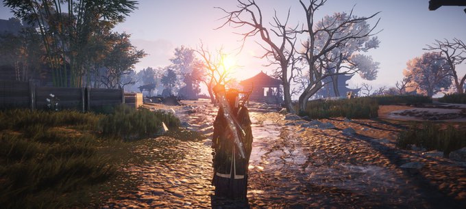 A scenic video game screenshot showing a character standing on a dirt path at sunset or sunrise, with trees and a small structure in the background.