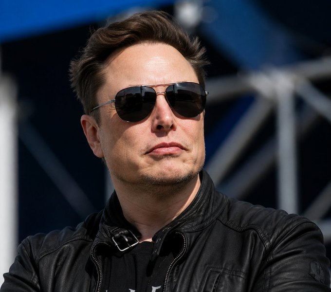 Elon Musk, wearing aviator sunglasses and a black leather jacket, looks out contemplatively at an outdoor event, his image often used to represent his leadership of X (formerly Twitter) and its ventures, including the platform's new crypto trading features mentioned in the accompanying post.