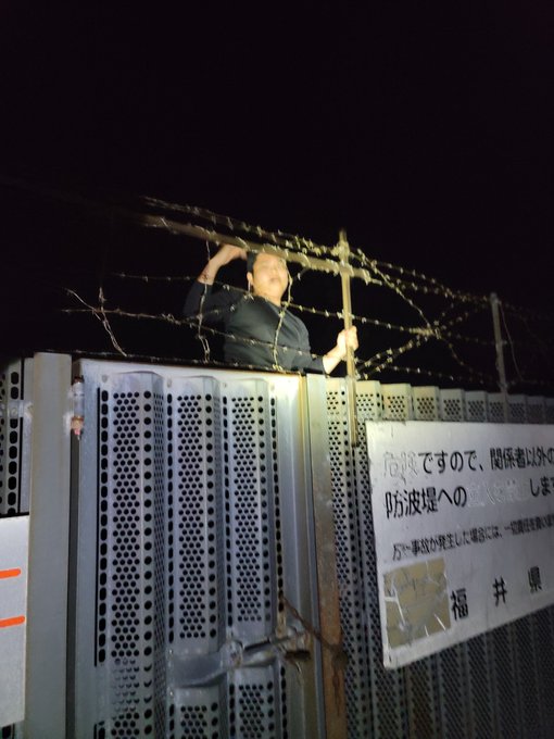 A person stands behind a barbed-wire-topped fence at night, gripping the bars, with a Japanese sign (including the characters for "Fukui Prefecture/福井県") visible on the perforated metal wall below. The staged "paparazzi" capture plays into the post's humor about being banned and reported for betcon, treating the playful "escape" as a sneaky nighttime photoshoot.