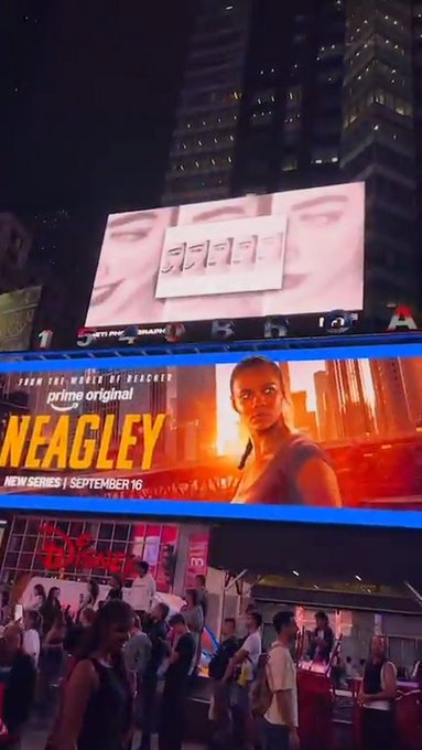 A nighttime city scene in a busy commercial district displays a large billboard advertising the Amazon Prime series 'REACHER' featuring the character 'NEAGLEY' with the release date 'NEW SERIES | SEPTEMBER 16'.